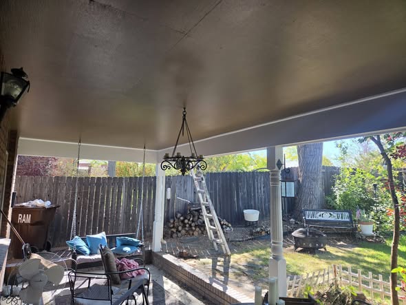 A covered patio ceiling being rolled with fresh paint, with a ladder in the yard behind